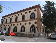 Edificio a due piani in stile storico con facciata intonacata chiara, bordi in mattoni e numerose finestre ad arco e oculi circolari con scuri scuri. Davanti c’è un parcheggio su strada e, sul lato destro, una scalinata esterna accanto ad alberi alti.