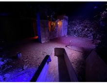 Area esterna notturna in un bosco con illuminazione blu e viola, con un percorso in terra e foglie. Al centro si vedono strutture sceniche in legno e pannelli a forma di rovine, con una luce calda sullo sfondo e un contenitore rettangolare in legno in primo piano.