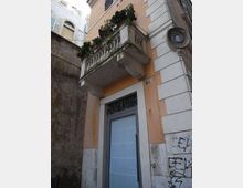 Facciata di un edificio urbano con balcone in muratura e balaustra, decorato con fioriere e vegetazione. Sotto il balcone si vede un portone alto con inferriata ornamentale, pareti intonacate e una targa rotonda sul muro laterale.