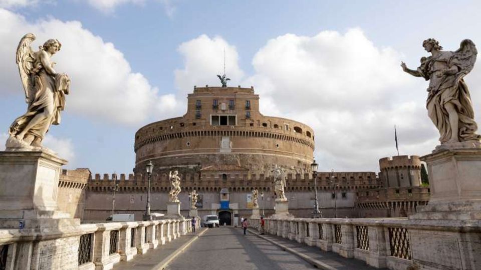 Veduta frontale di Castel Sant’Angelo con la sua struttura cilindrica in mattoni e le mura merlate, sullo sfondo di un cielo parzialmente nuvoloso. In primo piano il ponte con balaustre in pietra e statue di angeli scolpite lungo i lati conduce all’ingresso del castello.