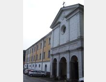 Facciata di un edificio religioso in pietra chiara con frontone e croce sulla sommità, rosone circolare e portico ad archi al piano terra con iscrizioni in rilievo. A sinistra si estende un’ala adiacente intonacata gialla con finestre ad arco, affacciata su una strada con auto parcheggiate.