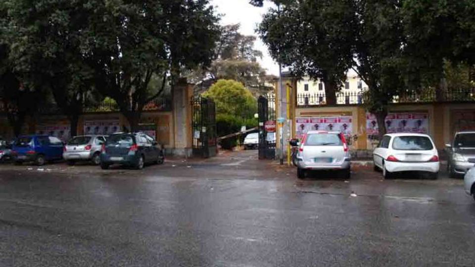 Ingresso con cancello in ferro tra pilastri in muratura, affacciato su una strada bagnata con auto parcheggiate ai lati. Grandi alberi ombreggiano l’area e un muro perimetrale con manifesti costeggia il lato destro.