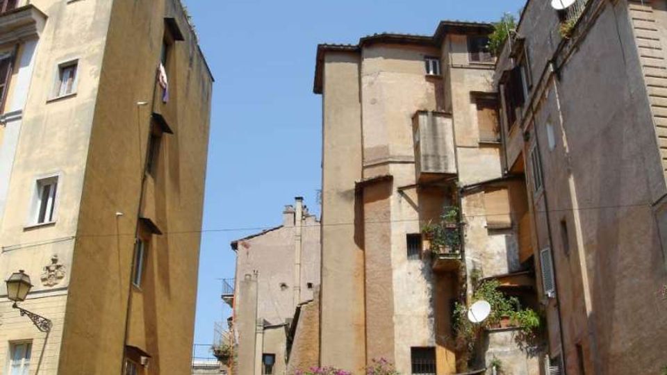 Cortile urbano tra alti edifici residenziali dai muri intonacati e usurati, con finestre e piccoli balconi irregolari. In basso si vede vegetazione con fiori e una lampada stradale, sotto un cielo azzurro.
