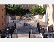 Ampia scalinata urbana terrazzata con rampe laterali e ringhiere metalliche, incassata tra alti edifici in mattoni. Muri bianchi con graffiti e alberi in cima incorniciano i diversi livelli collegati dalle scale.