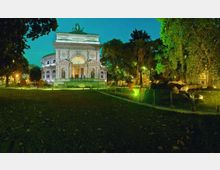 Edificio monumentale in stile classico illuminato di notte, con grande arco centrale e colonne, circondato da un parco alberato. In primo piano prato e percorso con illuminazione verde e recinzione bassa, con massi e vegetazione sul lato destro.