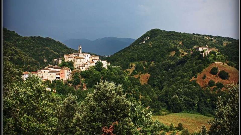 Veduta panoramica di un borgo arroccato con case in pietra e campanile, circondato da colline boscose e vallate verdi. Sullo sfondo si intravedono montagne e un cielo nuvoloso.