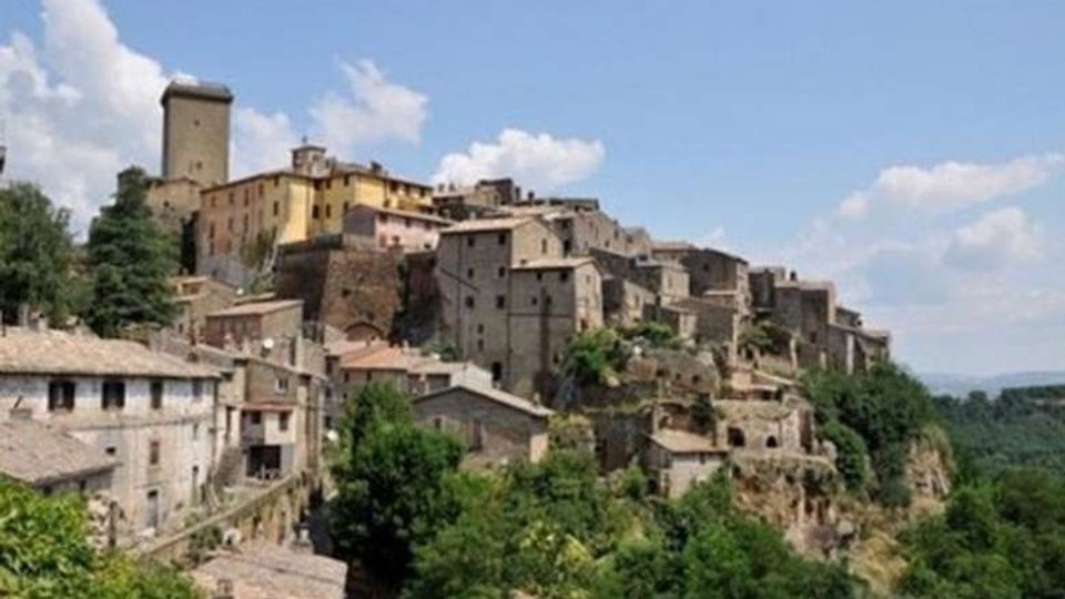 Borgo storico su un crinale con case in pietra addossate tra loro e una torre medievale prominente. Il complesso domina una valle verde con alberi e colline sullo sfondo sotto un cielo parzialmente nuvoloso.