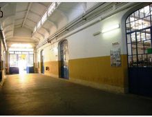 Corridoio interno di un edificio industriale con soffitto alto a volte, pareti bianche e fascia ocra, illuminazione al neon e tubazioni a vista lungo la parete. Grandi porte e finestre ad arco con telai metallici blu si aprono su altri ambienti e verso un ingresso luminoso in fondo.