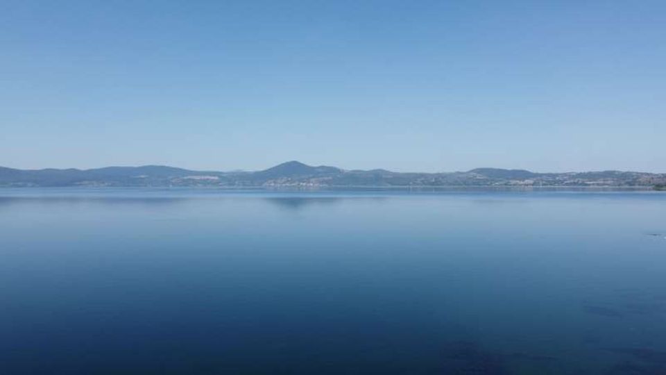 Vista panoramica di un grande lago con acqua calma e riflettente sotto un cielo sereno. All’orizzonte si vedono colline e una sponda lontana con edifici sparsi.