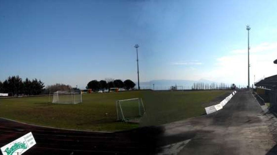 Campo da calcio all’aperto con pista di atletica in asfalto scuro, porte e piccoli tabelloni pubblicitari lungo il bordo. A destra si vedono tribune coperte, alti pali per l’illuminazione e una recinzione, con alberi e cielo sereno sullo sfondo.