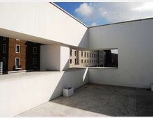 Terrazza in cemento di un edificio moderno, con alti muri bianchi e ampie aperture rettangolari che incorniciano la vista su palazzi residenziali vicini. Pavimentazione in lastre e cielo parzialmente nuvoloso sullo sfondo.