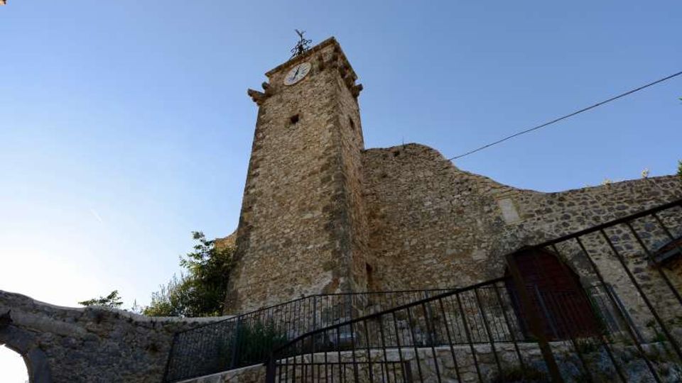 Torre campanaria in pietra di un edificio storico, con orologio vicino alla sommità e tetto a capanna collegato a un muro in muratura. In primo piano si vedono una scalinata in pietra e una ringhiera metallica, con cielo sereno sullo sfondo.