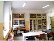 Aula adibita a biblioteca con armadi vetrati pieni di libri lungo le pareti e tavoli rettangolari con sedie in primo piano. Sul lato sinistro ci sono finestre e un banco con un computer, mentre il soffitto è illuminato da lampade fluorescenti.