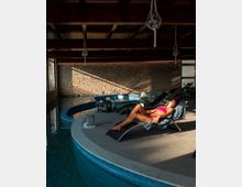 Area spa interna con piscina curva rivestita di mosaico blu, bordo in pietra e pavimento in piastrelle. Pareti in mattoni a vista, soffitto con travi in legno e lampade sospese, con lettini prendisole disposti lungo il perimetro.