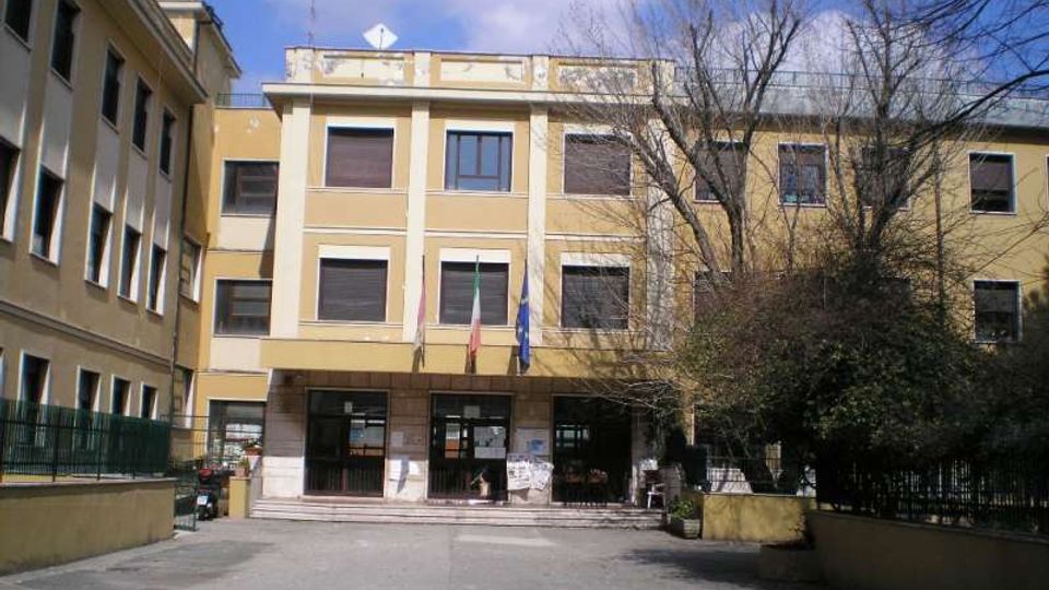 Edificio pubblico a tre piani con facciata gialla e colonne bianche, ingresso centrale con gradini e tre bandiere sopra la pensilina. Davanti si trova un cortile asfaltato, con recinzioni ai lati e alberi spogli sulla destra.