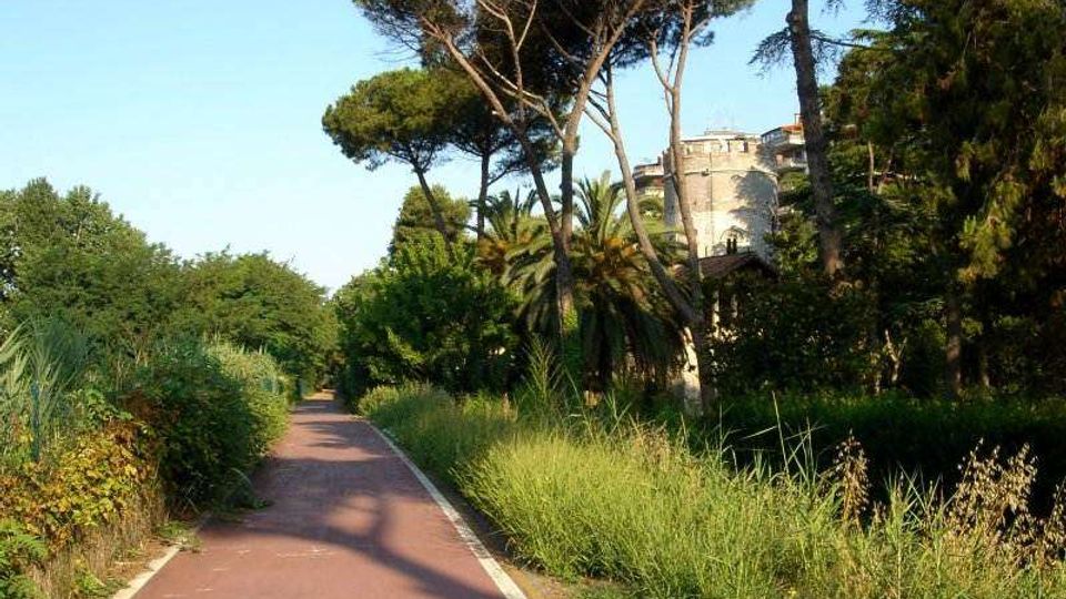 Pista ciclabile asfaltata che attraversa un’area verde con vegetazione mediterranea, cespugli e alti pini marittimi. Sullo sfondo si intravede un edificio in muratura parzialmente nascosto tra gli alberi.