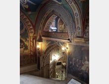 Interno di una chiesa o cappella con archi sovrapposti e soffitti a volta interamente decorati con affreschi e motivi geometrici. Una scala in pietra scende verso un livello inferiore illuminato da lampade a parete, con pavimento a mosaico e pannelli informativi.