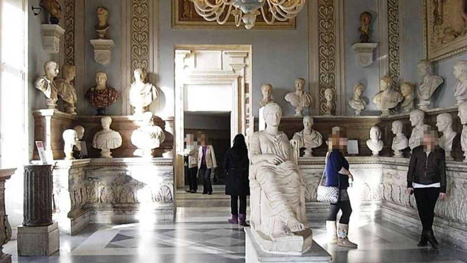 Sala museale classica con pareti ornate e cornicioni decorativi, illuminata da un grande lampadario in vetro e con pavimento in marmo a motivi geometrici. Busti e statue in marmo sono disposti su mensole lungo le pareti e una statua su piedistallo occupa il centro della stanza.