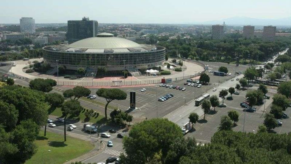 Veduta aerea di un grande edificio circolare con facciata vetrata e tetto a cupola, circondato da strade e ampi parcheggi. Intorno si estendono aree alberate e un contesto urbano con palazzi e grattacieli sullo sfondo.