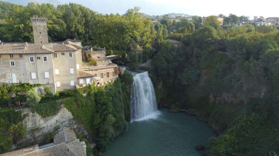 Veduta aerea di un borgo con edifici in pietra e una torre medievale affacciati su una gola boscosa. Al centro, una cascata scende in un bacino d’acqua verde circondato da pareti rocciose e vegetazione fitta.