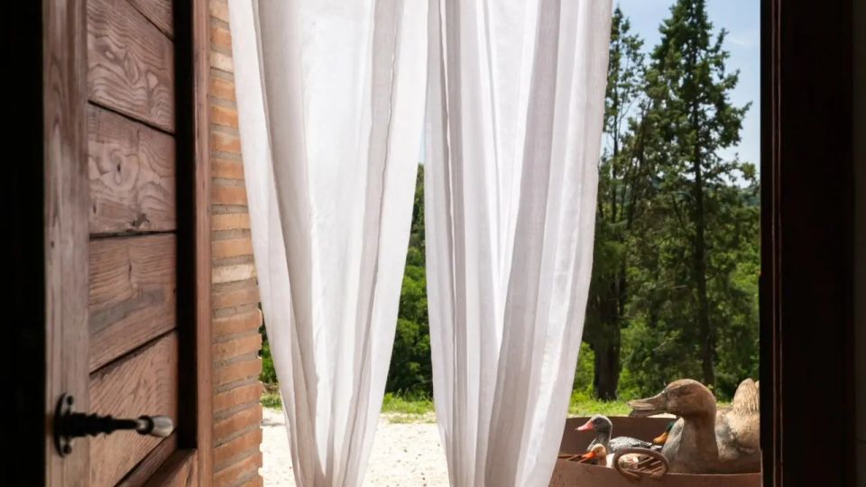 Porta in legno aperta su un patio in pietra, con tende bianche leggere che pendono e filtrano la luce. All’esterno si vede un cortile ghiaioso con un braciere su supporto metallico e alberi sullo sfondo sotto un cielo sereno.