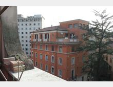 Veduta urbana dall’alto con un edificio in mattoni rossi a più piani, con finestre regolari e terrazze sul tetto con balaustre bianche. Sullo sfondo si vede un alto palazzo bianco e, sul lato destro, un grande albero sempreverde vicino agli edifici.