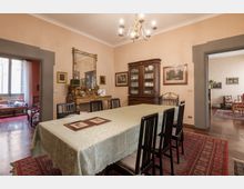 Sala da pranzo in stile classico con grande tavolo rettangolare coperto da tovaglia chiara, sedie scure e tappeti orientali su pavimento in legno. Alle pareti ci sono quadri e uno specchio sopra una consolle decorata, con lampadario a bracci sul soffitto e vetrina in legno; due ampie aperture collegano la stanza ad ambienti adiacenti.