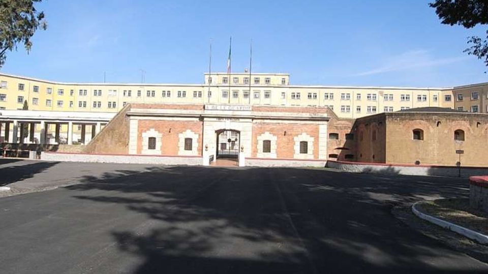 Ampio piazzale asfaltato davanti a un complesso architettonico con ingresso centrale in mattoni e pietra, con cancello ad arco e finestre piccole. Sullo sfondo si vede un grande edificio istituzionale giallo a più piani, con facciata simmetrica e una bandiera sul tetto.