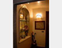 Ingresso interno con nicchia ad arco in pietra e mensole in vetro che espongono oggetti decorativi. Pareti color crema con applique a parete, quadro con mappa incorniciata e porta scura; a terra un portaombrelli con ombrelli.