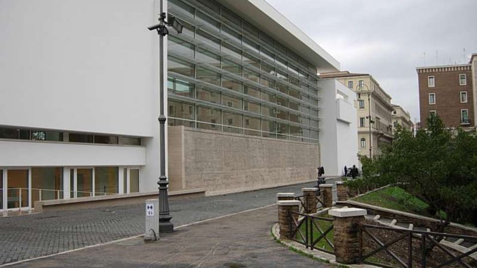 Edificio moderno con ampia facciata vetrata a pannelli orizzontali e volumi bianchi, affacciato su una strada pavimentata con lampione. In primo piano si vede un vialetto curvo con parapetto in pietra e legno che costeggia un’area verde, con palazzi urbani sullo sfondo.