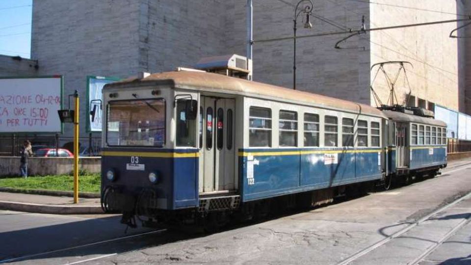 Tram blu e crema su rotaie in una strada urbana, con cavi elettrici aerei e palo della linea. Sullo sfondo si vedono grandi edifici in cemento e un marciapiede con segnaletica e cartelloni.
