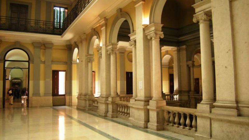 Ampio cortile interno di un edificio storico con pavimento in marmo lucido, porticato con alte colonne in pietra e grandi archi. Al livello superiore corre una balconata con ringhiera in ferro battuto, con finestre e porte lungo le pareti color ocra.