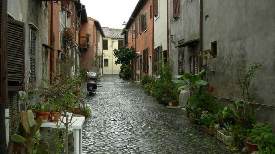 Vicolo stretto in un borgo con pavimentazione in ciottoli, fiancheggiato da case a due piani con facciate intonacate e persiane in legno. Lungo i muri ci sono numerosi vasi e fioriere con piante, e in fondo si intravede un edificio chiaro con tetto in coppi.