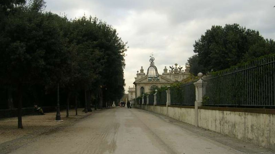 Lungo viale alberato con strada sterrata che conduce a un edificio monumentale sullo sfondo, con facciata classica decorata da statue e una cupola centrale. A destra corre un alto muro con ringhiera in ferro, mentre a sinistra ci sono alberi e lampioni lungo il margine del percorso.