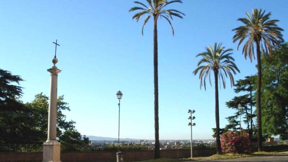 Belvedere con piazzale lastricato e vista panoramica sulla città in lontananza, delimitato da un muretto basso. In primo piano ci sono alte palme, un lampione e una colonna in pietra sormontata da una croce.