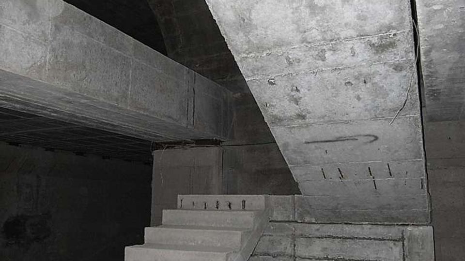 Interno in cemento grezzo di un edificio o bunker sotterraneo, con una scala in calcestruzzo che sale verso un solaio inclinato. Travi e pareti massicce in cemento a vista creano un ambiente angusto e poco illuminato.