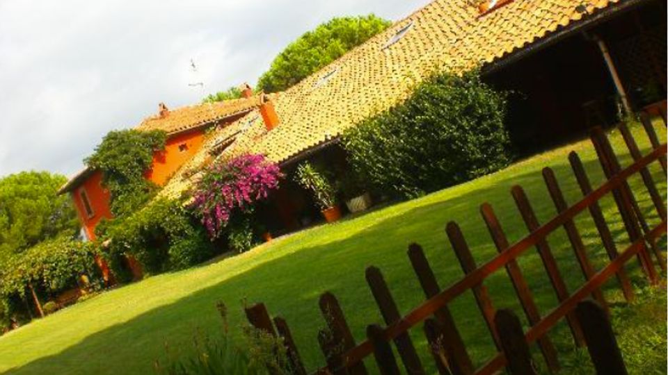 Casa di campagna con tetto in tegole e facciata arancione, circondata da un ampio prato verde e cespugli fioriti. In primo piano si vede una staccionata in legno lungo il bordo del giardino.