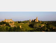 Panorama di una collina verde con resti di un castello e mura in pietra, con una torre cilindrica e altri ruderi distribuiti lungo il crinale. Sullo sfondo si estende la campagna con alberi e campi sotto un cielo limpido.