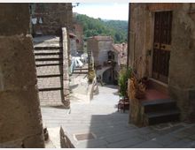 Vicolo in pietra in un borgo collinare, con scalinata e case in muratura ai lati. La strada scende verso un piccolo arco tra gli edifici, con tetti in coppi e colline boscose sullo sfondo.