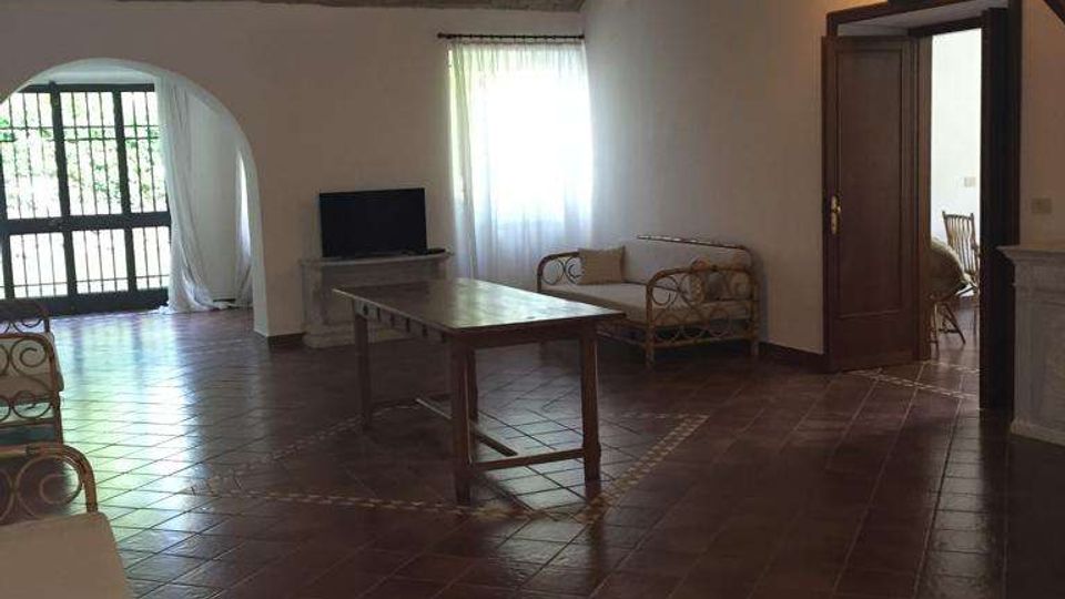 Ampio soggiorno rustico con pavimento in cotto, soffitto con travi in legno a vista e pareti bianche. Al centro c’è un tavolo, con divanetti in rattan, una finestra con tenda e una grande porta ad arco vetrata che dà sull’esterno.