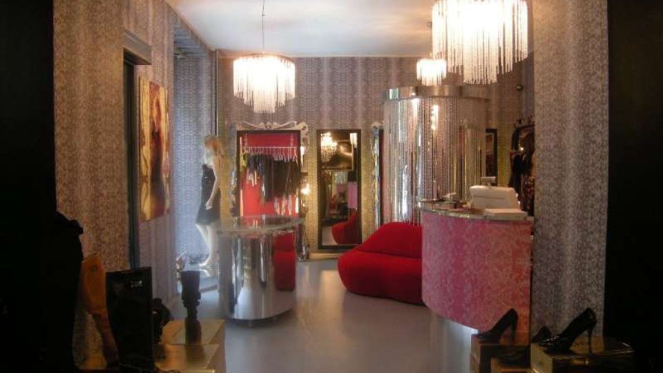 Interno di una boutique di moda con pareti rivestite da carta da parati, lampadari a frange pendenti e pavimento grigio. Lo spazio include un bancone curvo rivestito in tessuto rosa, un divano rosso, specchi e espositori con scarpe e abbigliamento.