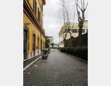 Strada acciottolata in un cortile urbano, con un lungo edificio giallo con persiane e portoni sulla sinistra e un edificio chiaro sulla destra. Lungo il percorso ci sono grandi vasi con piante, una fila di alberi spogli e siepi vicino a un muro, sotto un cielo nuvoloso.