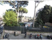 Parcheggio urbano con pavimentazione in sanpietrini e una fila di scooter lungo un muro in mattoni con recinzione metallica e cancello. Dietro la recinzione si vedono alti pini e altre alberature, con edifici e tetti della città sullo sfondo.