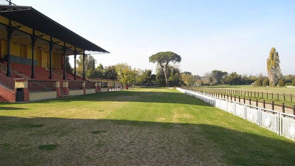Vista di un campo erboso lungo una tribuna coperta con struttura in metallo e gradinate in muratura. A destra corre una lunga recinzione bianca che delimita un’area aperta, con alberi e vegetazione sullo sfondo sotto cielo sereno.