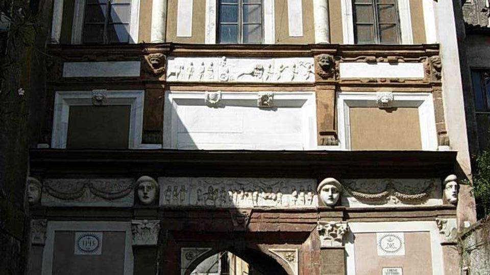 Facciata di un edificio storico con decorazioni in stucco e medaglioni, finestre alte incorniciate e una grande arcata centrale che funge da passaggio verso una strada stretta. Alla base si vede un portale ad arco in pietra con bassorilievi e cartelli stradali accanto, in un contesto urbano tra edifici ravvicinati.