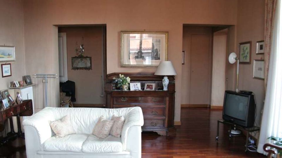 Soggiorno domestico con pavimento in parquet e pareti color rosa tenue, arredato con un divano bianco, una cassettiera in legno con cornici e lampada, e un televisore su supporto nell’angolo. Due aperture interne conducono ad altre stanze e quadri incorniciati decorano le pareti.
