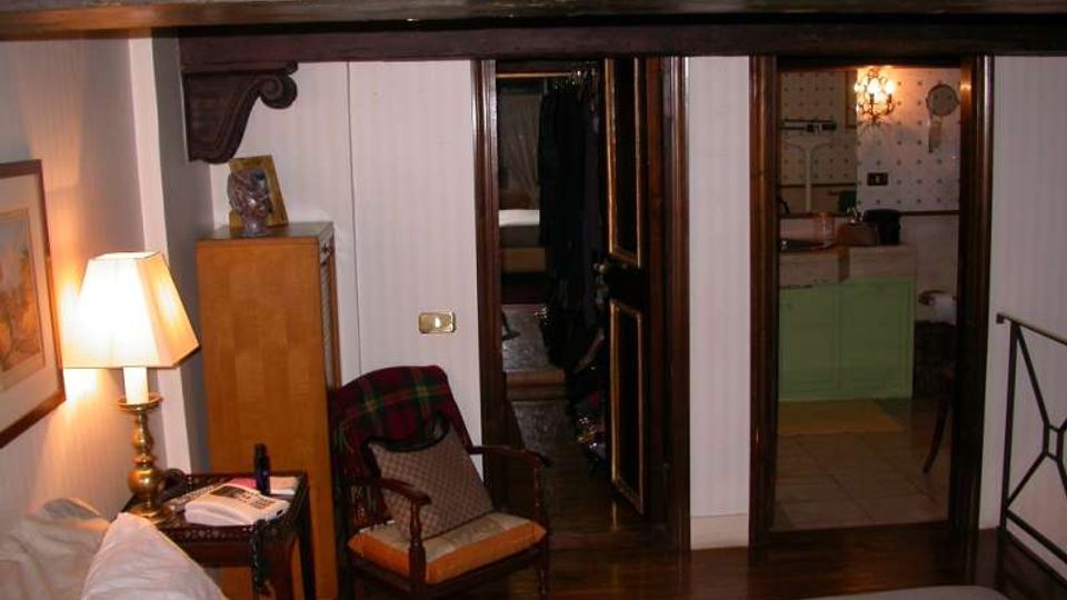 Interno di una camera da letto con soffitto a travi e arredi in legno, comprensivo di letto in primo piano, comodino con lampada e poltrona. Sullo sfondo si vede un corridoio con porte in legno che conduce a un bagno con piastrelle chiare e mobile lavabo verde.
