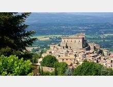 Veduta panoramica di un borgo collinare con case in pietra dai tetti in coppi, dominato da un grande castello-fortezza rettangolare con torri. Sullo sfondo si estende una valle con campi e boschi, con colline e montagne lontane.