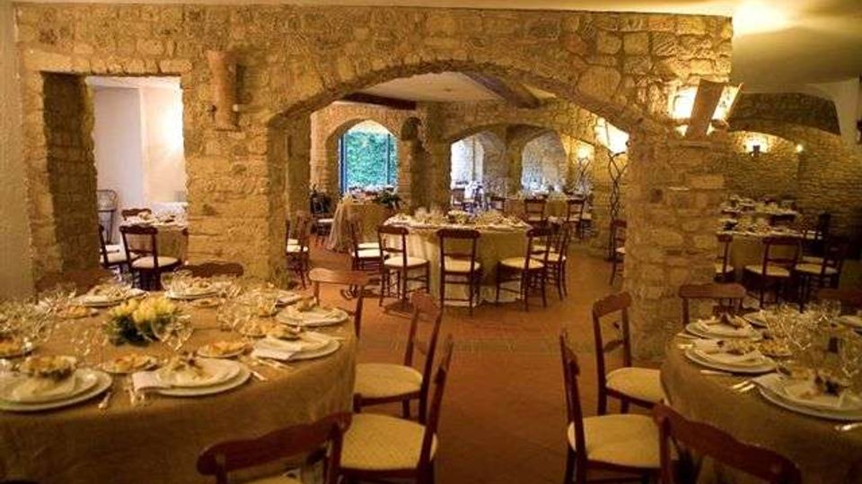 Sala da pranzo in stile rustico con grandi archi e pareti in pietra a vista, pavimento in terracotta e illuminazione calda. Tavoli rotondi apparecchiati con tovaglie e sedie in legno disposti in più aree sotto le arcate.
