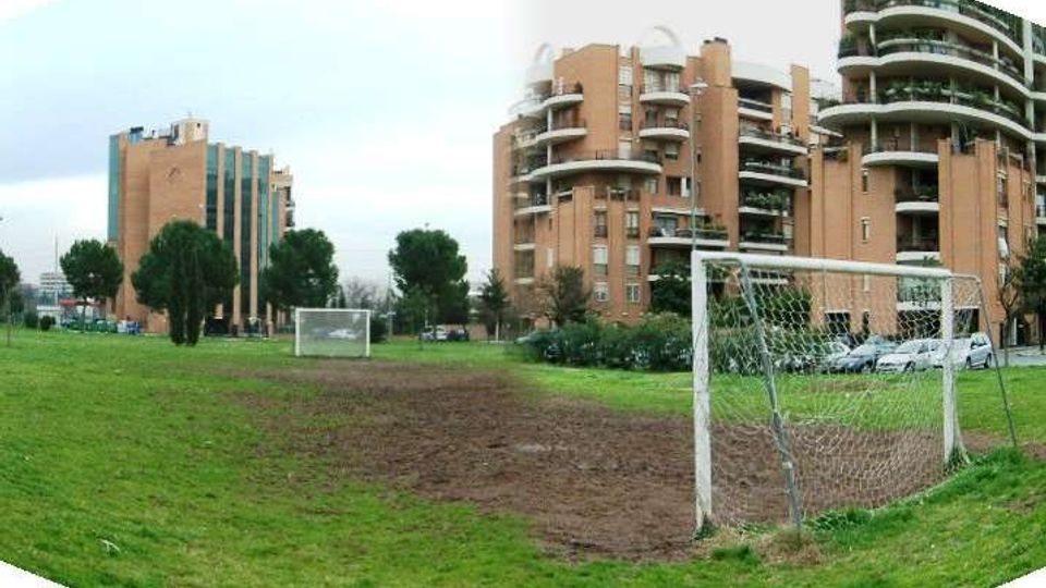 Campo da calcio in erba con aree di terra battuta e due porte, situato in un parco urbano. Sullo sfondo si vedono alti edifici residenziali in mattoni con balconi curvi, alberi e auto parcheggiate.